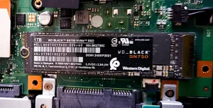 A green motherboard with a black chip on top of it. On the left side a screw keeps the chip in place. The text on the chip reads 1 TB, WD Black™ SN 750 NVMe™ SSD. It has the Western Digital logomark. It's not clear if the chip is connected or not, as it seems to be just placed flat over the motherboard.