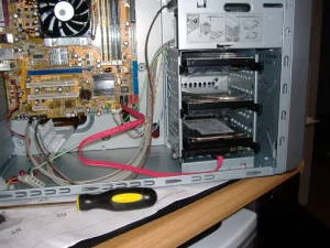 An open PC case. In it a large orange electronic board can be seen. On its bottim-right corner, 2 black ports and 2 red ports can be see. On the black port a red, flat cable is connected. The other end of the cable is connected to a metallic device in a device inside the PC, behind the front panel. Above the device similar devices can be seen stacked one on top of the other.