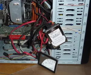 A PC case opened, various cables coming out of it. Thick black cables and red flat cables connect to two devices hanging out of the PC by the cables. The first device is a thick metallic rectangular block that reads "Toshiba." The second is flatter and has the Intel logomark.