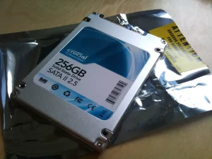A rectangular, blocky device. Connects at its top. On it, written: 256 GB Solid-State Drive, SATA II 2.5. The Crucial logomark can be seen. Under it a black plastic bag slightly larger than the device.