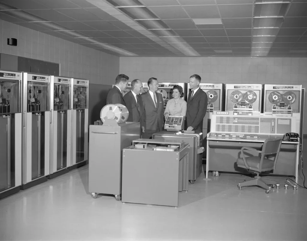 A black and white photograph featuring five people talking in a room. Next to the walls, devices as tall as the people can be seen. Each with two circular shapes inside a glass pane that are larger than a person's head. A chair in front of a panel full of buttons can also be seen.